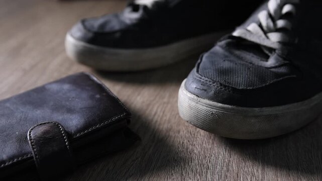 Poor financial situation. Putting black wallet near old sneakers onto wooden floor, closeup