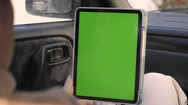 Person holds tablet with green screen inside car. Focus on device, not person's face. Ideal for mockups, apps, or digital content. Car interior suggests travel or road usage. Suitable for advertising