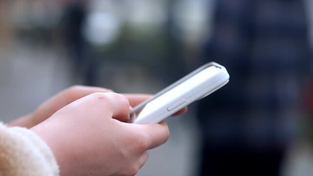 Person holds smartphone outdoors, fingers typing. Background blurred, focus on hands and device. Neutral expression, intent on communication or browsing. Suitable for social media, tech