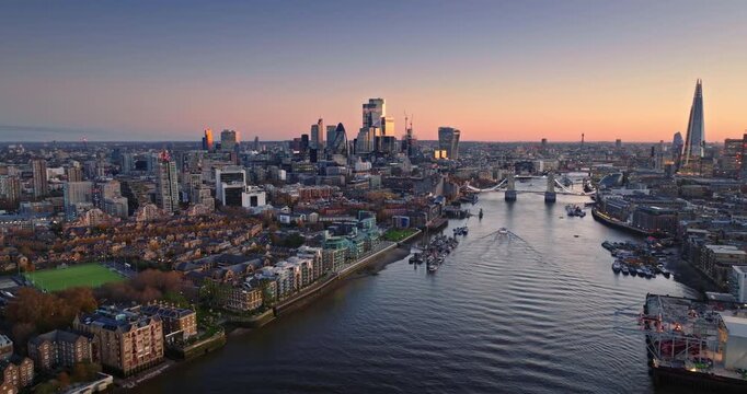 London city financial district skyline rising alongside the River Thames as a boat creates a wake on the water while the sun sets over the urban landscape. Evening cityscape aerial view drone panorama