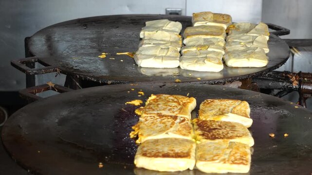 A folded Martabak Telor Kubang pastry filled with seasoned eggs, minced beef, onions, and aromatic spices is carefully placed onto a large hot griddle for cooking. 