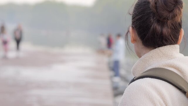 Person walks alone on wet pavement, hair tied up. Backpack strap visible, coat warm and cozy. Blurred background shows others walking in distance. Mood calm, reflective, peaceful