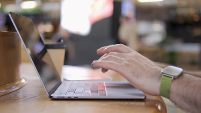 Hands type on laptop in cozy cafe setting. Warm lighting enhances relaxed, focused atmosphere. Person wears smartwatch with green band. Ideal for remote work, freelancing, or digital creativity scenes