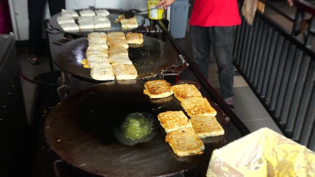 A folded Martabak Telor Kubang pastry filled with seasoned eggs, minced beef, onions, and aromatic spices is carefully placed onto a large hot griddle for cooking. 