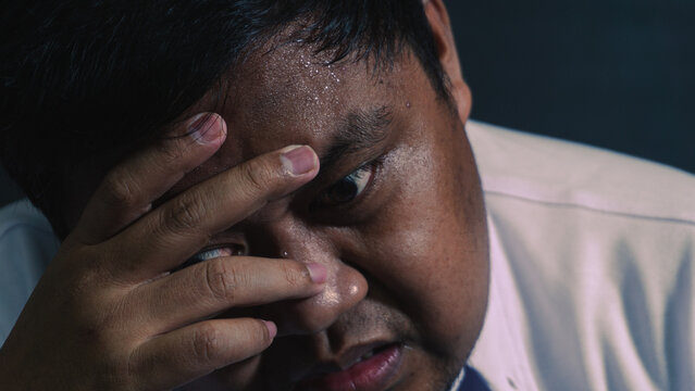 An intense macro portrait shows an exhausted adult Asian male covering the face with one hand in deep sorrow. The heavily sweating skin and wide staring eye convey a terrifying panic