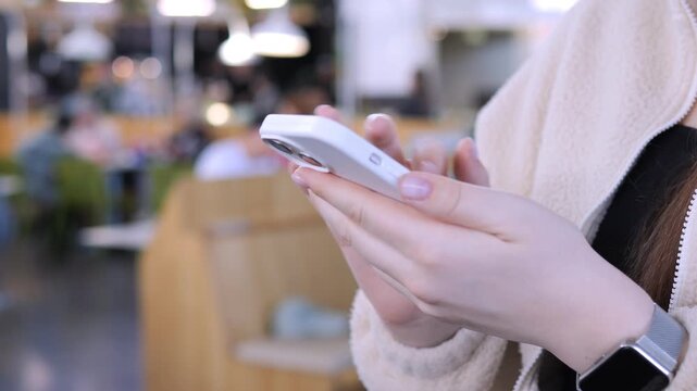 Close-up of hands holding smartphone. Person wears cozy sweater and smartwatch. Background shows blurred cafe with wooden furniture. Focus on interaction with device. Suitable for tech, communication