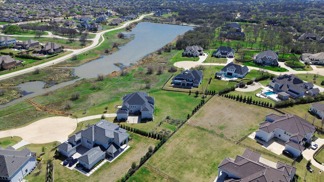 Retention pond sits central to custom home developments within Dallas suburb Tour 18 subdivision. High angle view reveals cul de sacs, sprawling lawns, dark shingle roofs. Flower Mound, TX
