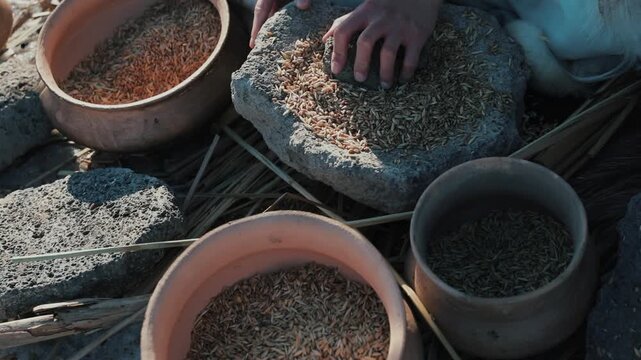 Detailed close-up of hands grinding grain using an ancient stone mill and clay pots, demonstrating primitive food preparation.