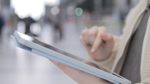Person holds tablet, taps screen. Hand gestures show interaction. Background blurred with moving people. Modern device use in public space. Urban setting with soft natural lighting
