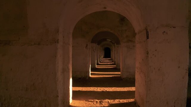 Cinematic shadows and shafts of sunlight moving across the floor of a dark, historic arched underground corridor in an ancient redoubt.
