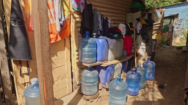 Outdoor storage of multiple blue water jugs arranged in rows next to a house wall with clothing hung up above.