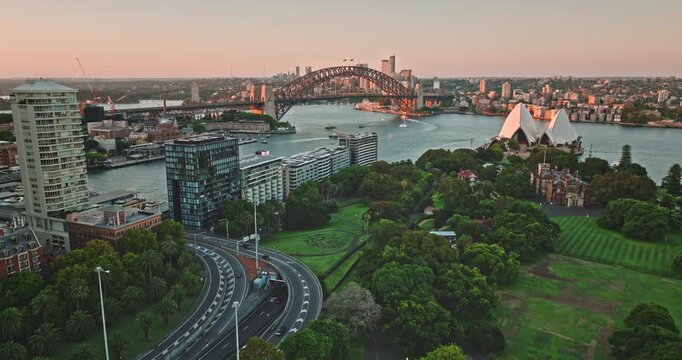 Aerial drone flight over famous Sydney landmarks, the iconic Opera House and Harbour Bridge, standing prominently along the vibrant harbour, framed by city buildings and green parkland at golden hour