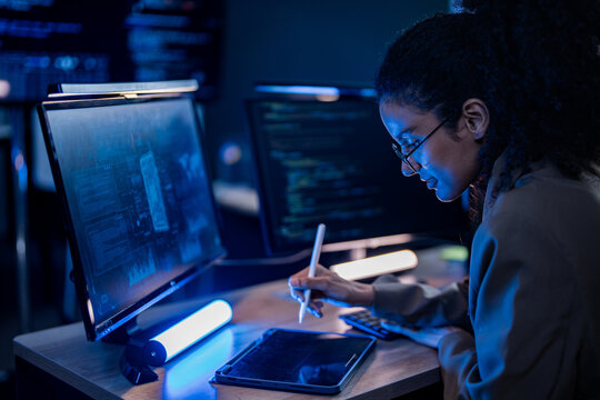 A female developer reviews software output on a glowing display during a focused late-night session at her desk, in a spacious, dimly lit modern research facility.