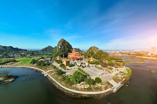 Bright orange temple buildings stand on a peaceful green peninsula near the river in Da Nang under a vibrant morning sky with tall limestone cliffs nearby
