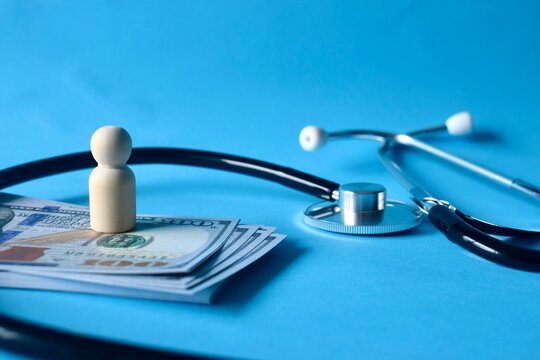 A high-angle view of a stack of hundred-dollar bills with a wooden pawn figure and a stethoscope on a blue background representing health insurance costs.
