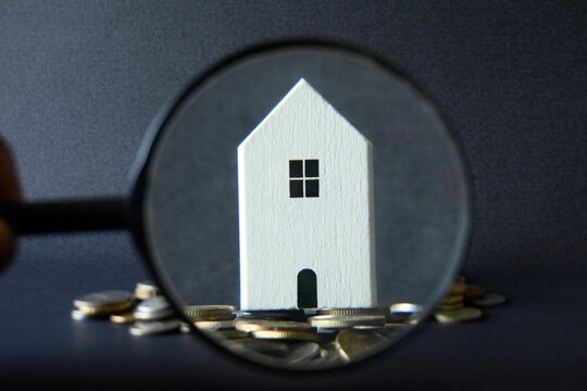 A conceptual financial photo showing a detailed white miniature house focused sharply through a magnifying glass held over a pile of coins.