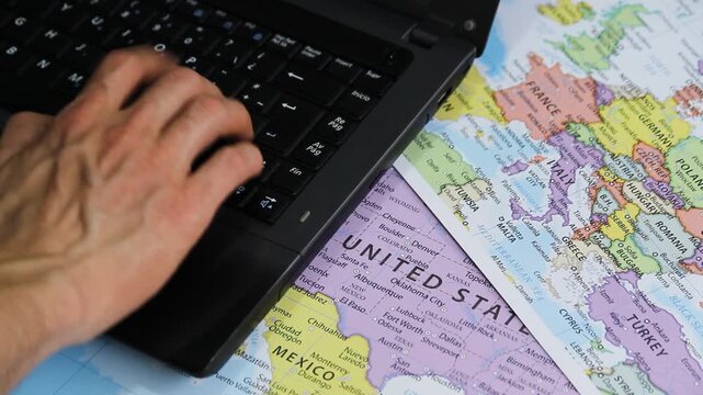 Detailed view of a male hand typing on a black laptop with a politic world maps at background in a travel concept
