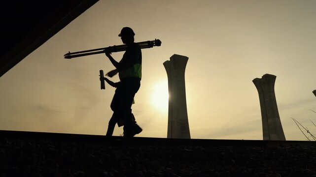 Silhouette of a survey engineer carrying a tripod across a construction site during sunset with concrete bridge columns in the background, symbolizing engineering, infrastructure, and fieldwork.
