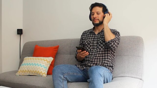 A latin man at the right side of the screen sitting on a couch listening music on a smartphone with a headcase