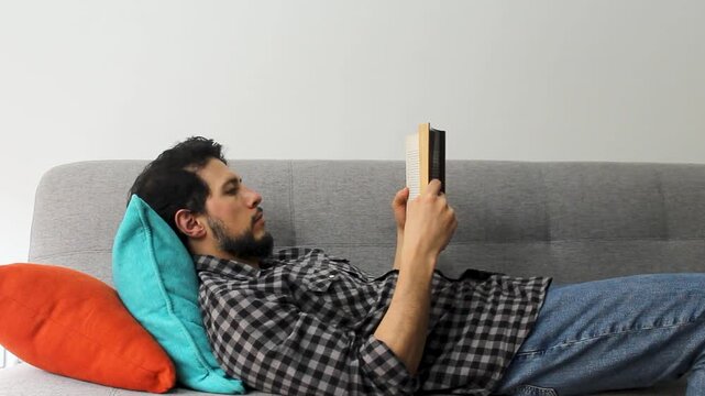 A hispanic man with beard lying on a couch reading a text book in a peaceful attitude