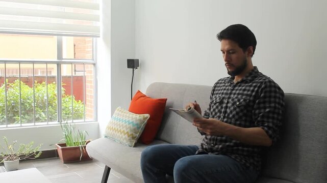 A hispanic man sitting on a couch of a living room solving words puzzles in a concentrating attitude