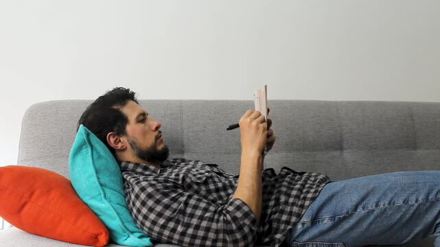 A latin man with beard lying on a couch solving crosswords puzzles in a peaceful attitude
