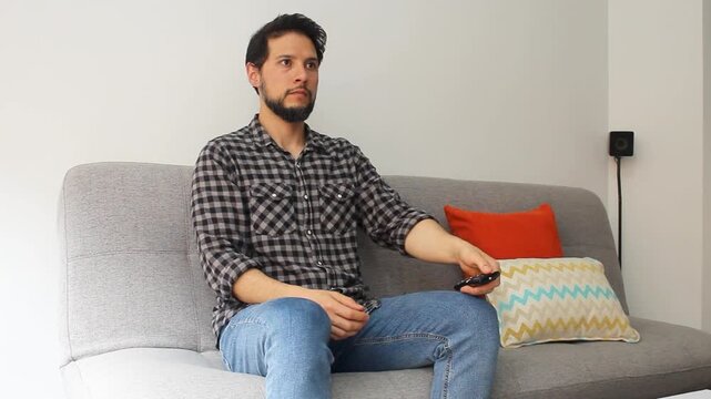 A latin man sitting on a couch turning on the TV with a remote controller and choosing options to see