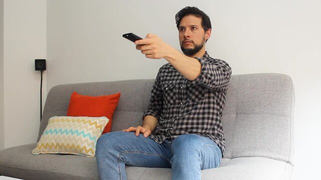 A latin man sitting on a couch at the right side of the screen using a remote controller of a smart tv