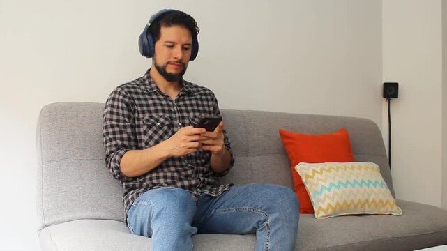 A hispanic man sitting on a couch using a wireless headset texting in his smartphone in a happy attitude