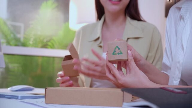 Closeup hand two asian woman holding cardboard box with recycle for eco friendly packaging design, team businesswoman working and meeting showing paper product prototype in modern office.
