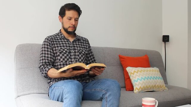 A caucasic hispanic man reading a text book sitting at the left side of a couch in a concentrating attitude