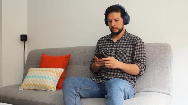 A latin man sitting at the right side of a couch using a wireless headset texting in his smartphone