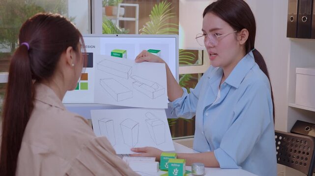 Two asian businesswoman working packaging design with ai computer, team business woman meeting of paper sketch drawing mockup holding prototype product box, creative packaging design project.