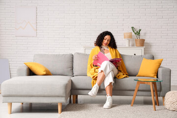 Beautiful young African-American woman with cozy blanket reading fashion magazine on sofa at home © Pixel-Shot