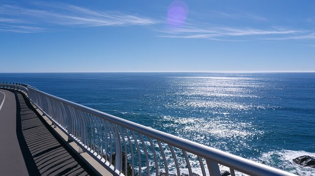 Scenic outdoor coastal drive landscape, gently curving grey road on left, long white metal guardrail separating road and vibrant blue ocean extending to horizon, sunlight shimmering on soft waves, bri