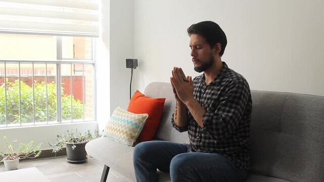 A caucasic man with a rosary bead on his hands praying with his eyes closed sitting on a couch in a concentrating attitude