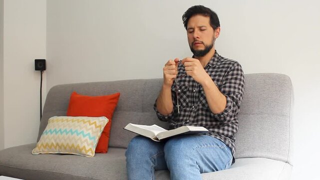 Detailed view of a latin man with his eyes closed at the right side of the couch praying the rosary with a holy bible on his legs