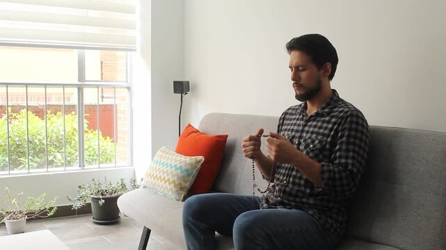 a latin man with his eyes closed praying the rosary in concentrating attitude sitting on a couch