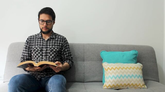 Detailed view of a latin male with eyeglases sitting at the left side of a couch reading a text book in a concentrating attitude