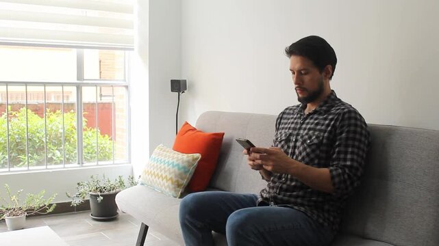 A latin man sitting on a grey couch typing on his smart phone in a peaceful attitude