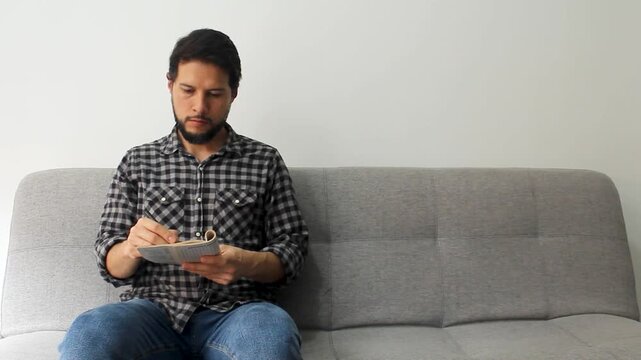 Detailed view of a caucas man sitting at the left side of a grey couch solving crosswords puzzles in a concentrated attitude