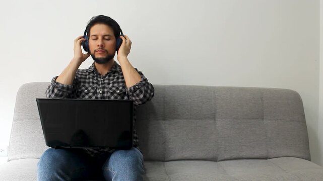 Detailed view of a happy man with his eyes closed sitting at the left side of a grey couch listen music on a wireless headset with a laptop over his legs