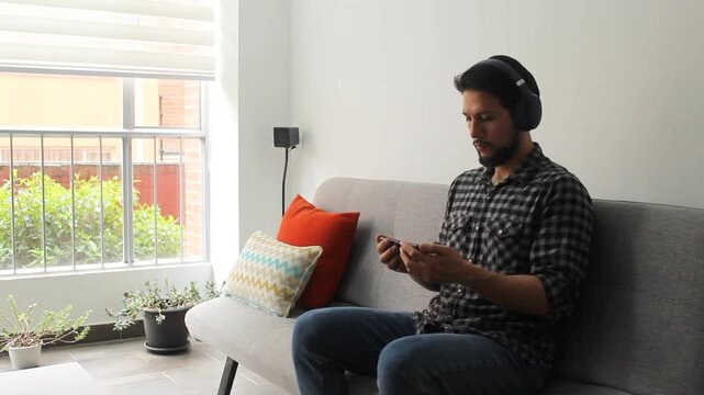 A caucasic male sitting on a grey couch watching something on a smartphone with a wireless headset in a concentrating attitude