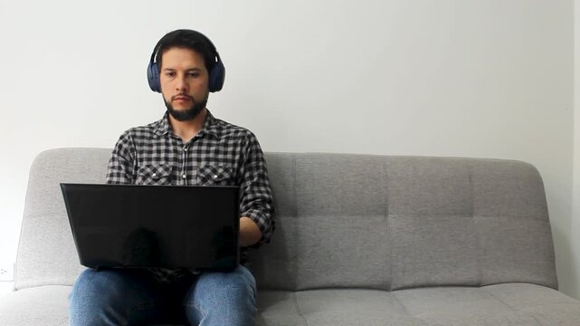 Detailed view of a latin man sitting on the left side of a couch working remotely on a black laptop over his legs in a concentrated attitude