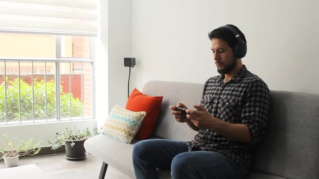 A latin man playing games on his phone sitting on a grey couch in a living room