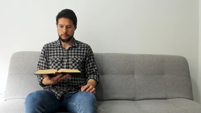 Detailed view of a male sitting at the left side of a grey couch reading the holy bible in a concentrating attitude