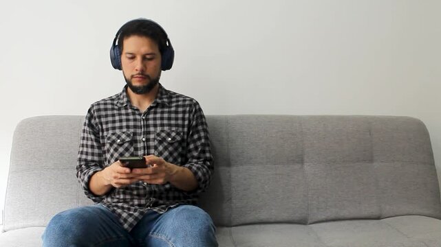 Detailed view of a male sitting at the left side of a couch typing on a smartphone and using a wireless headset