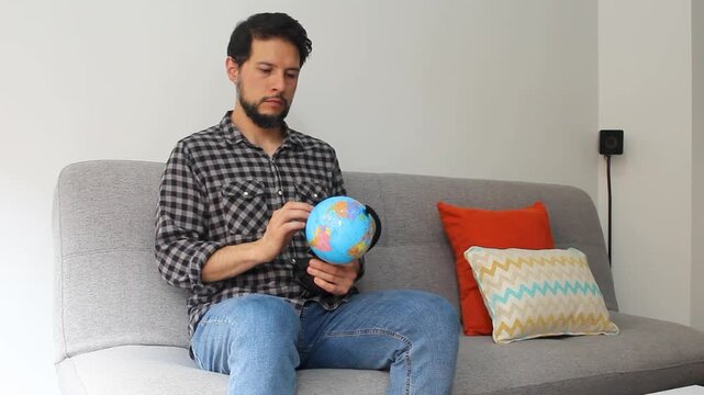 Detailed view of a latin man sitting at the left side of a grey couch spinning with his hand a world globle in a concentrating attitude