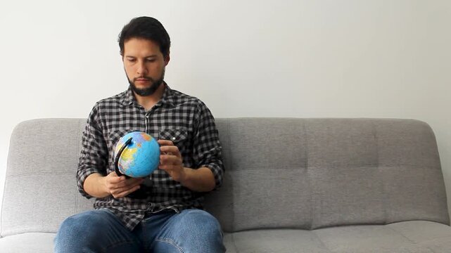 Detailed view of a latin male sitting on a grey couch spinning with his hand a politic world globe in a travel concept