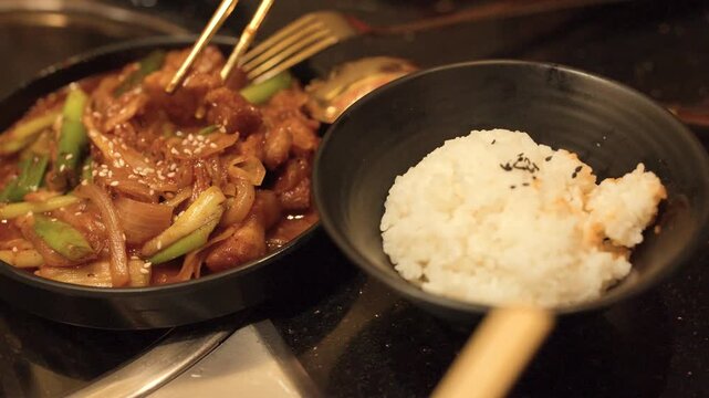 Chopsticks Placing Spicy Pork Bulgogi onto White Rice in Black Bowl